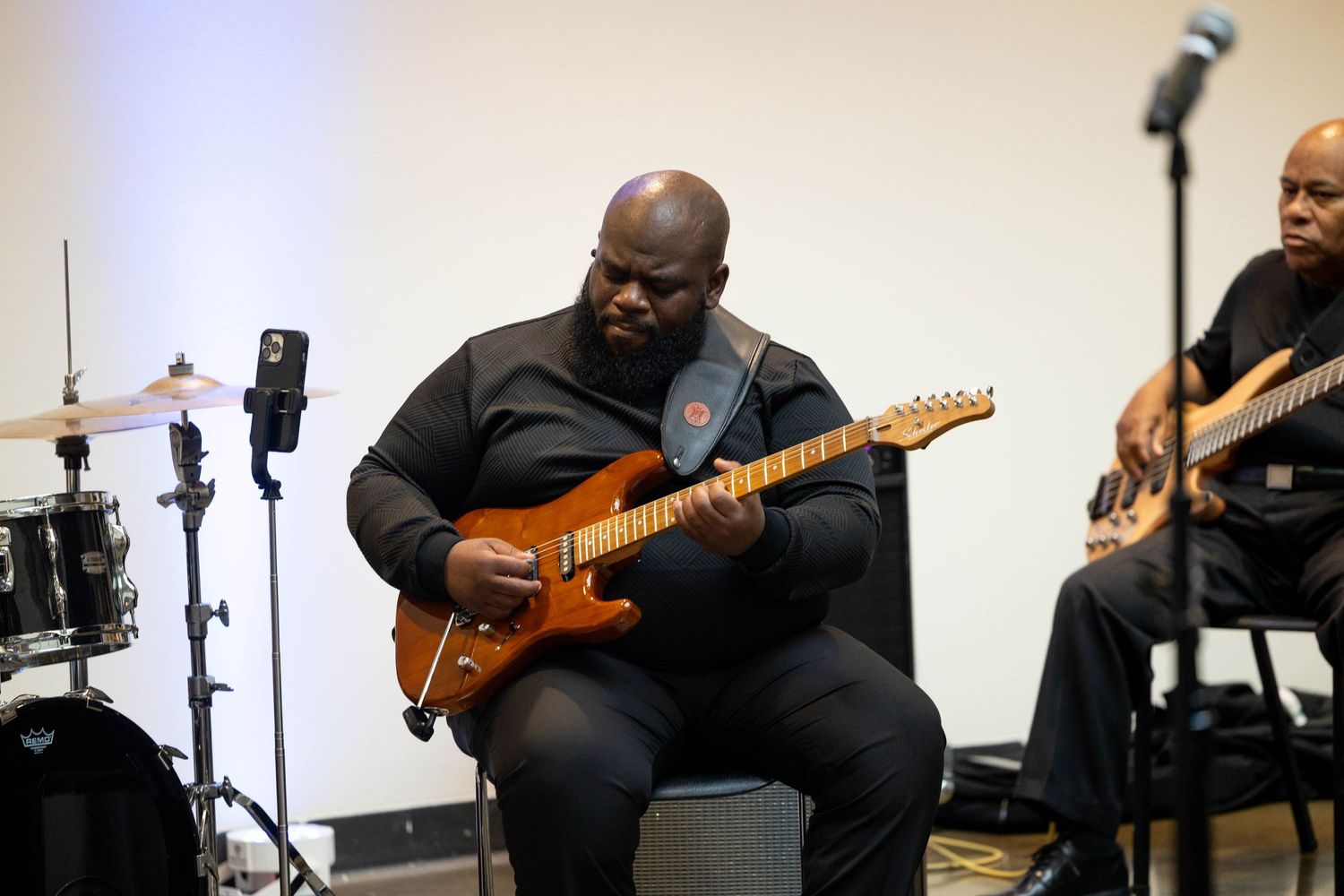 Musician playing electric guitar on stage during live performance with drummer and bassist in background.