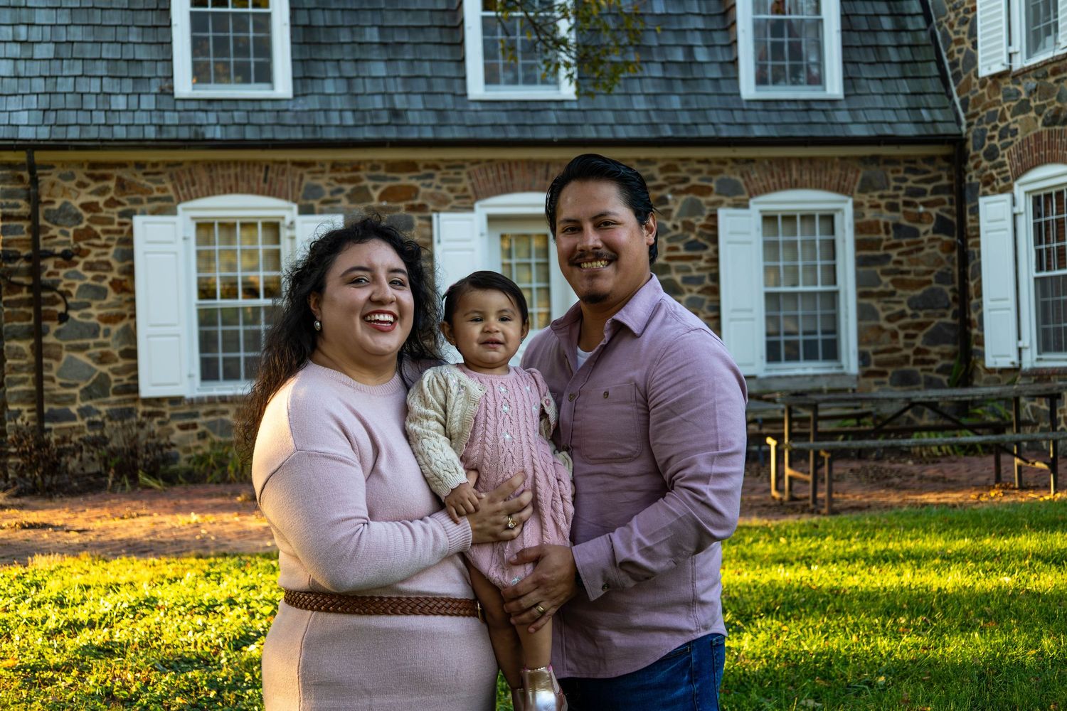 Happy family of three posing outdoors, parents holding baby girl, smiling in front of a stone building.