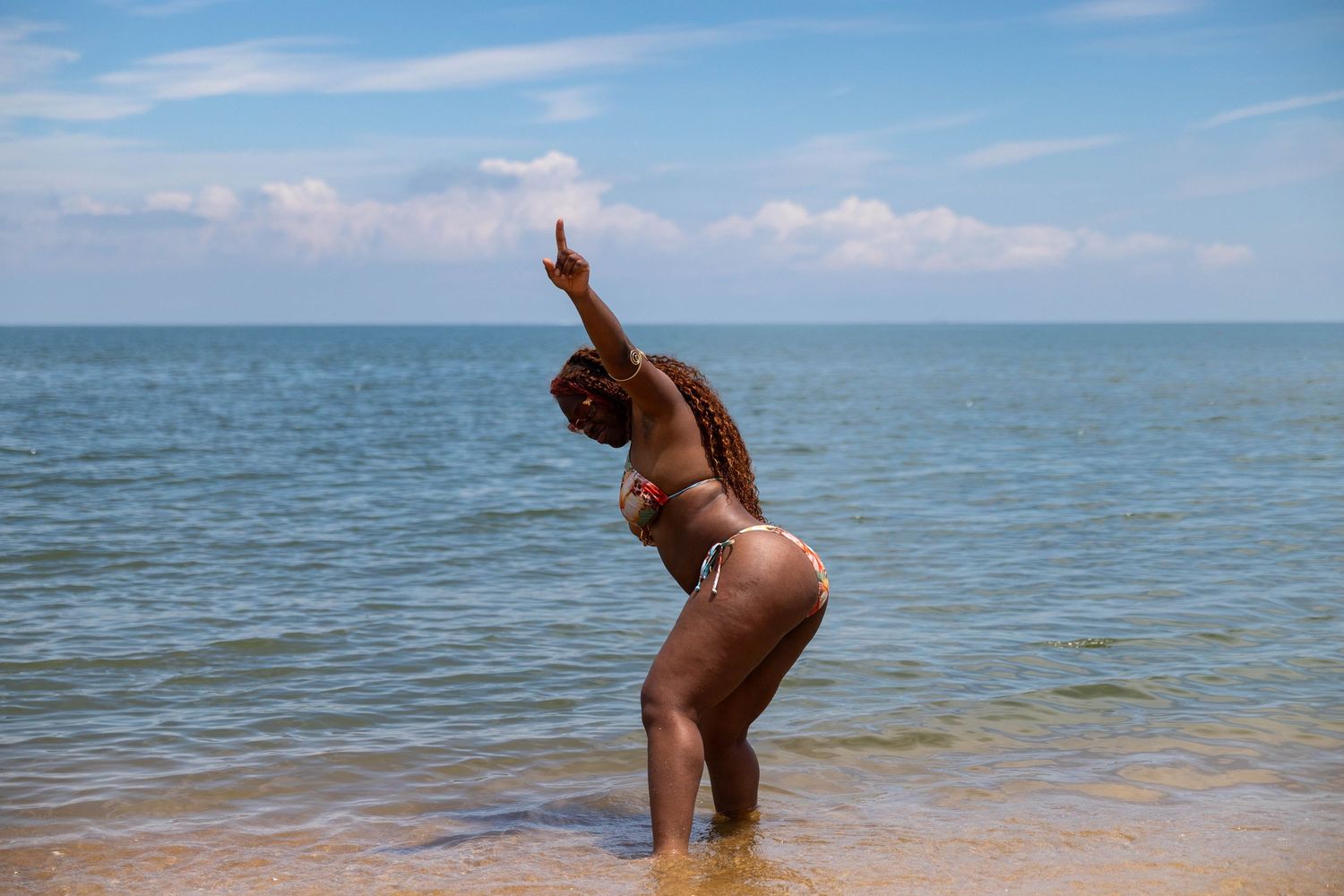 Woman in bikini posing with arm raised at the beach with calm blue ocean waters behind her.