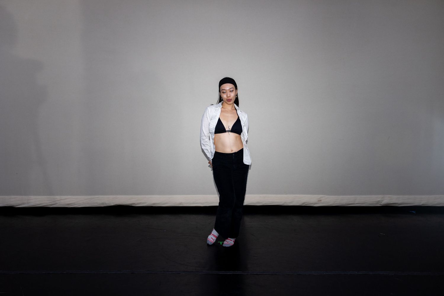 Woman in black bralette and wide-leg pants wearing white jacket posing in a studio.
