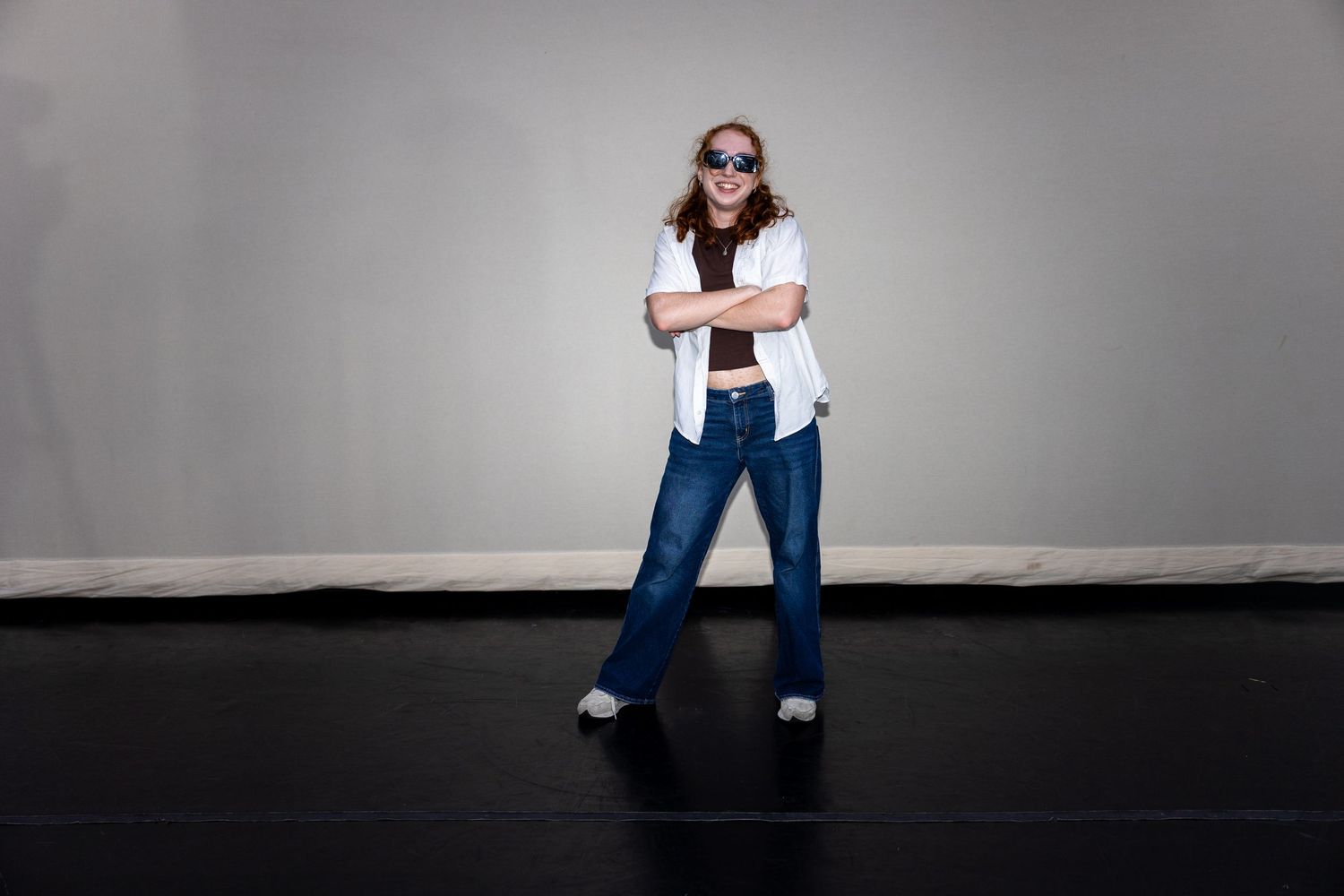 Woman in white shirt, jeans, and sunglasses standing with arms crossed in studio.