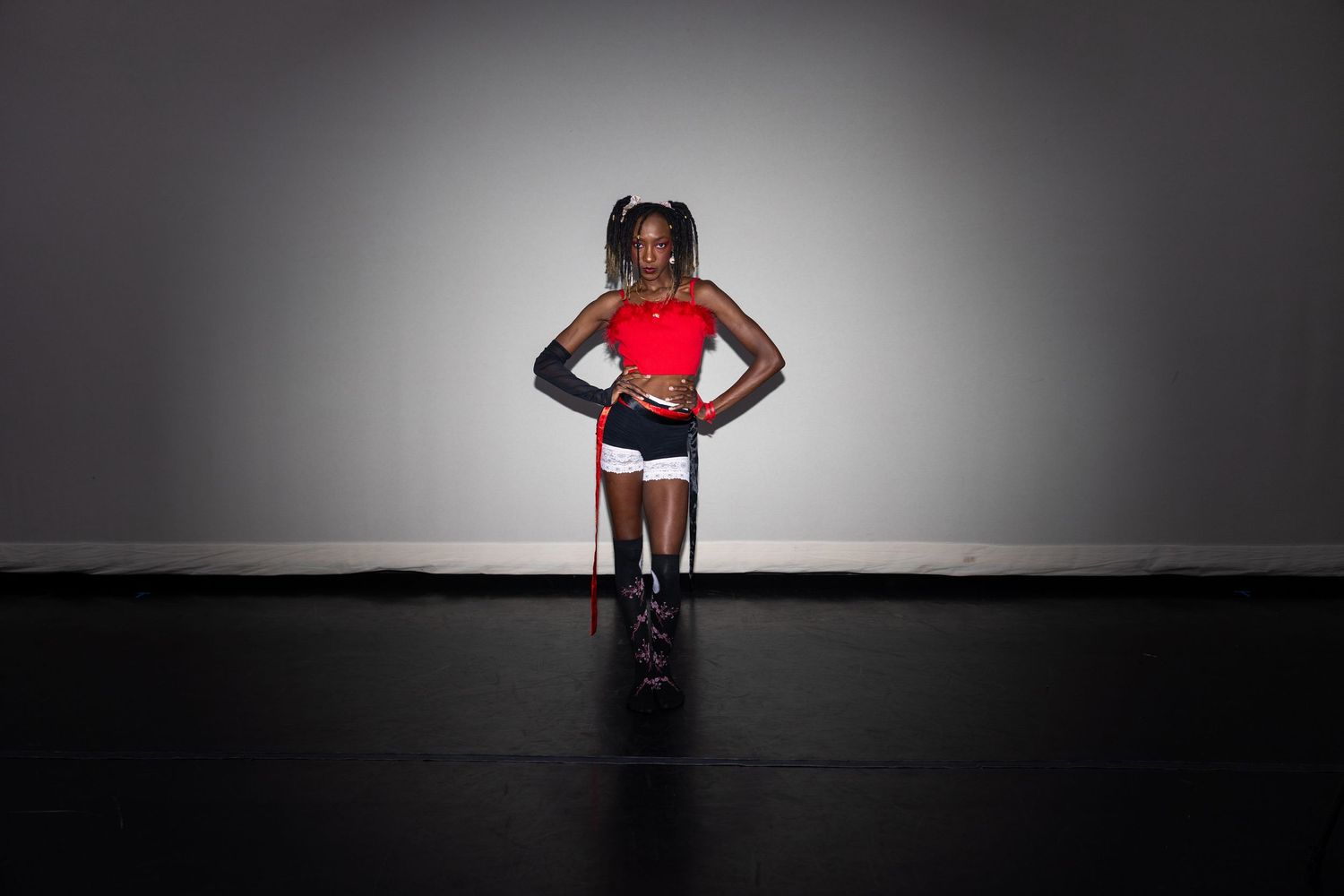 Young Black female dancer in red crop top and white shorts poses confidently on dark stage floor.