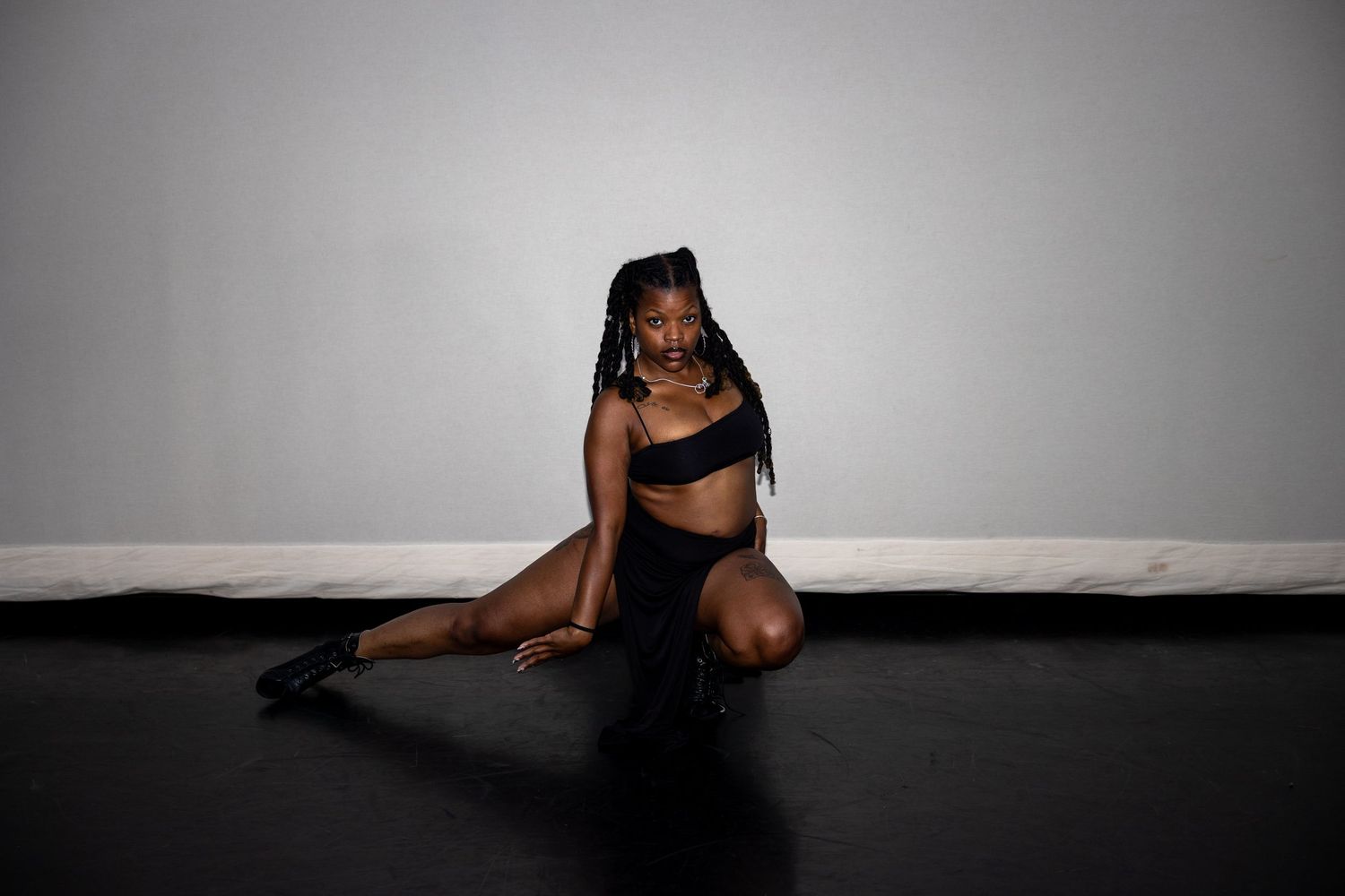A dancer in black bralette and shorts sits elegantly on a dark stage floor in a studio.