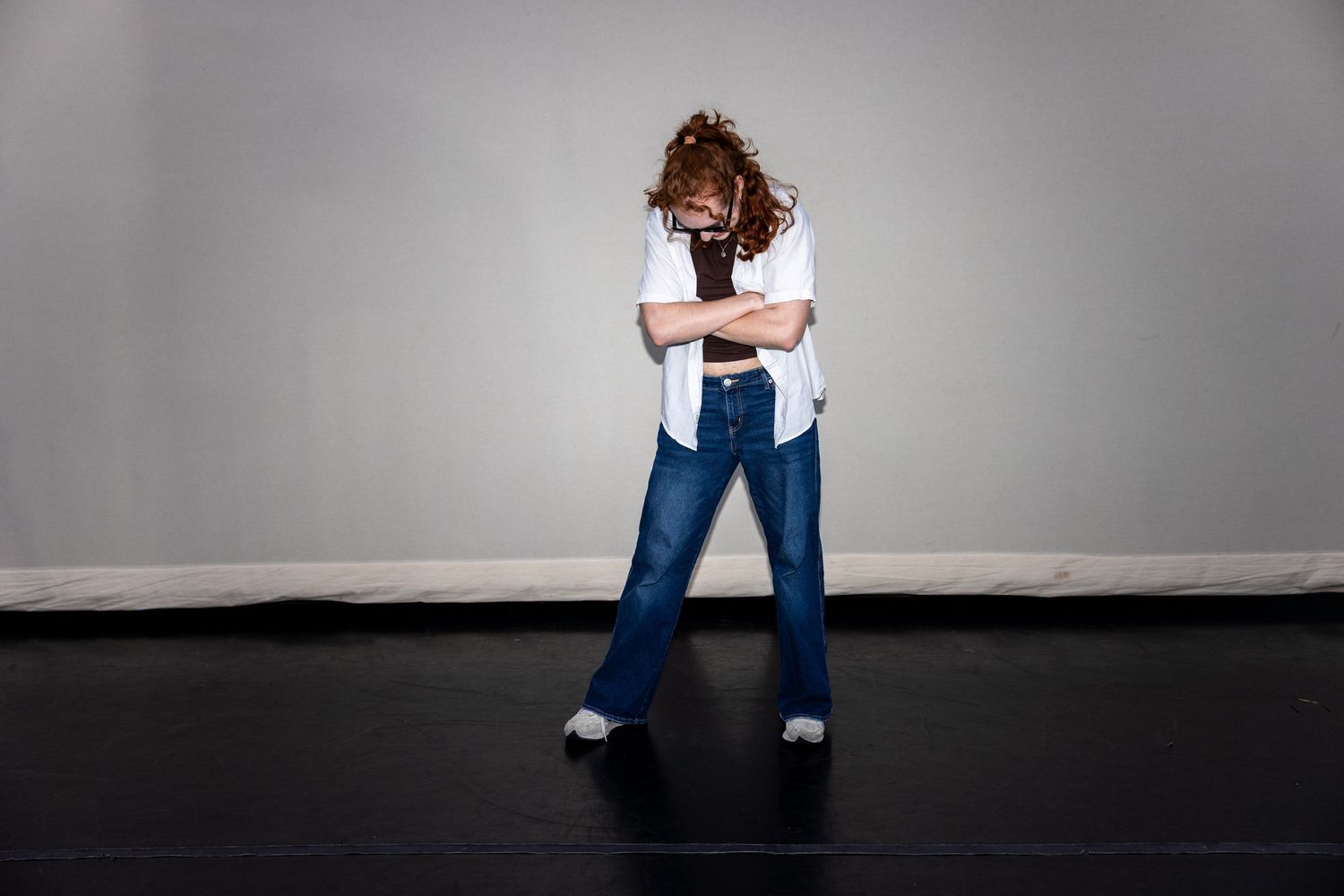 Curly-haired dancer stands pensively in white shirt and blue flare jeans on black stage.