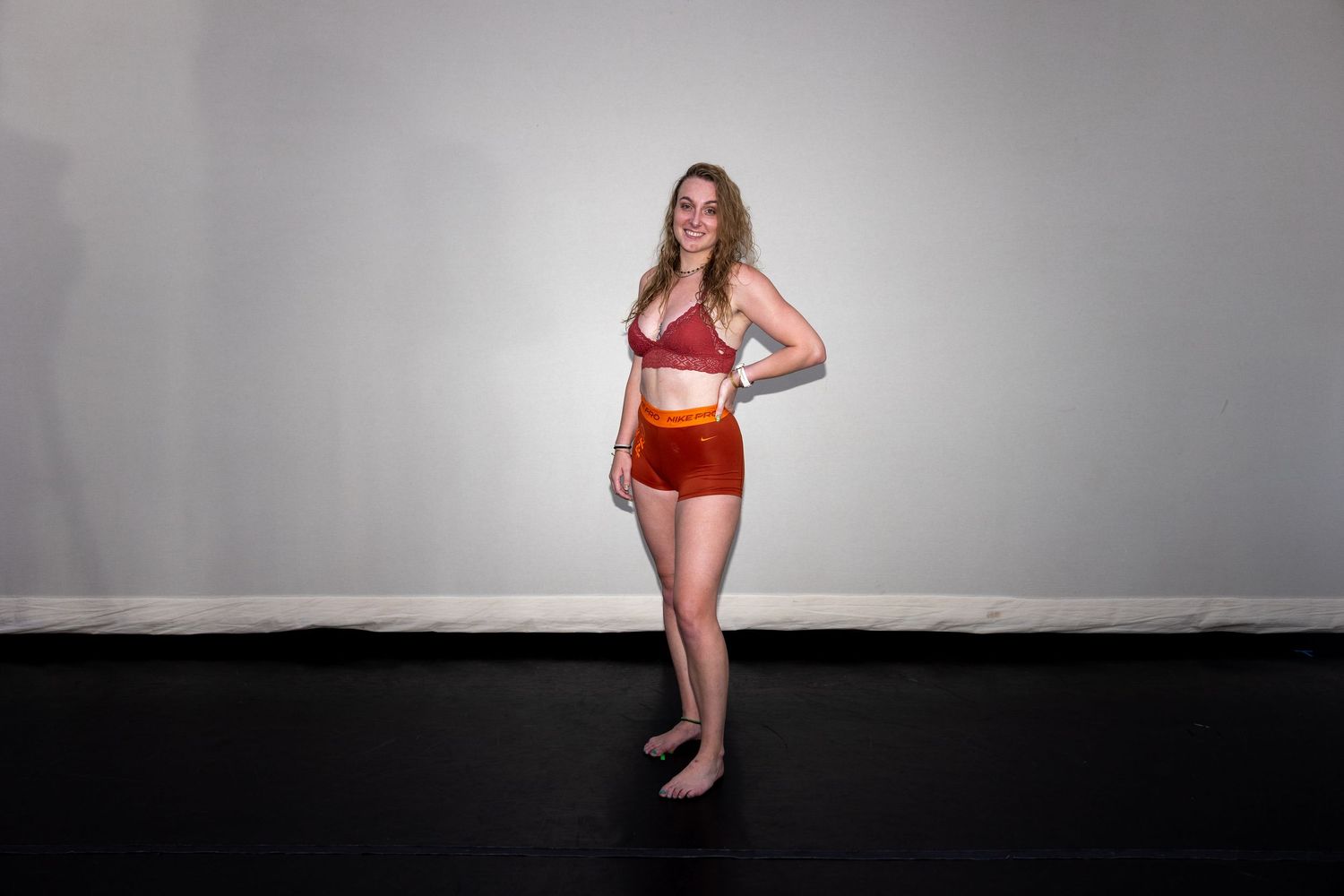 A woman in a red sports bra and orange shorts stands barefoot against a white studio wall.