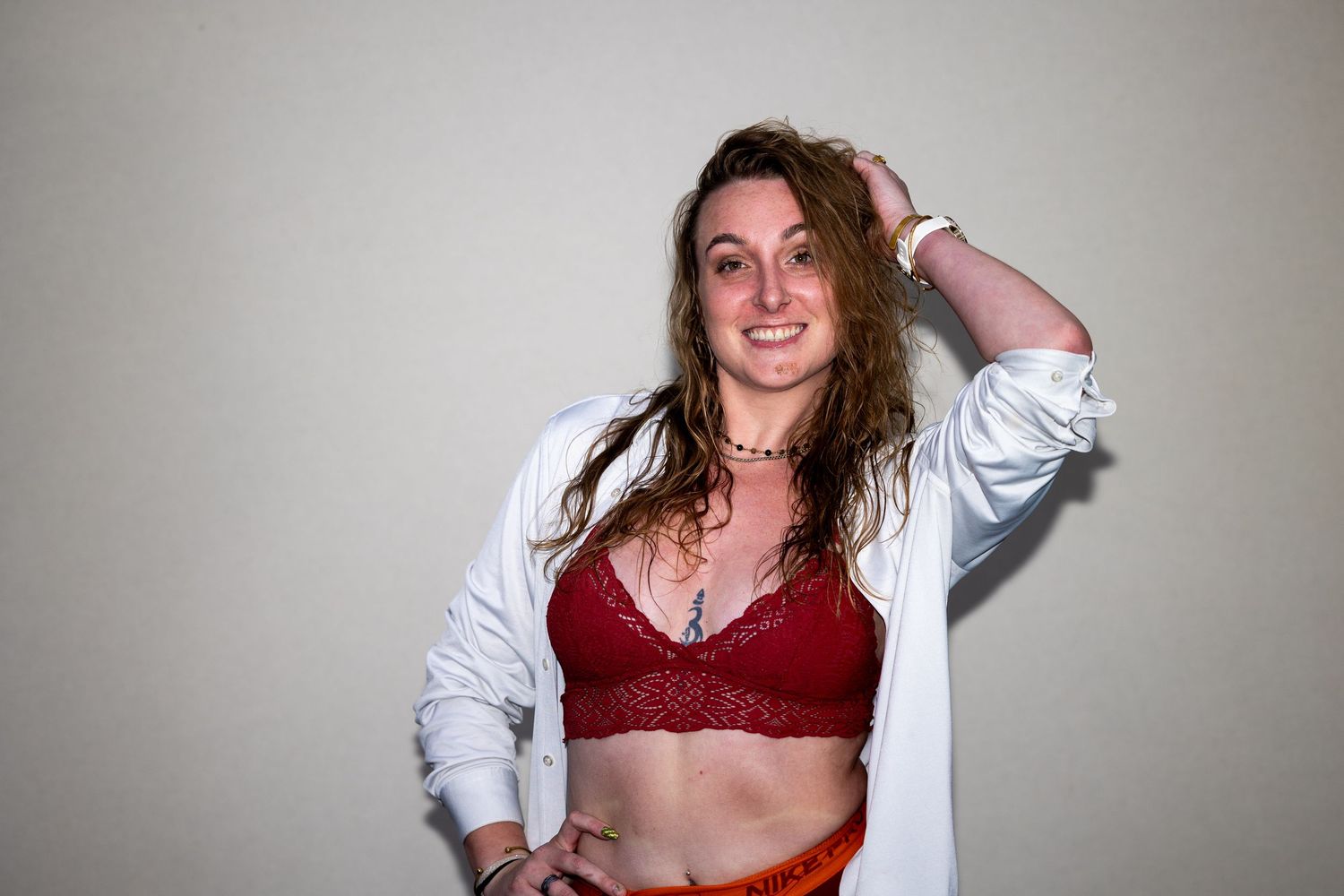 A woman in a red lace bralette and white open shirt smiles while posing with hand in her hair.