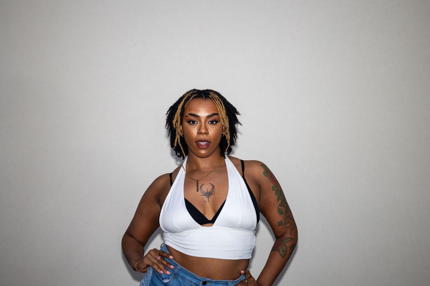 Confident woman with braids in white halter crop top and jeans with tattoos posing against white background.
