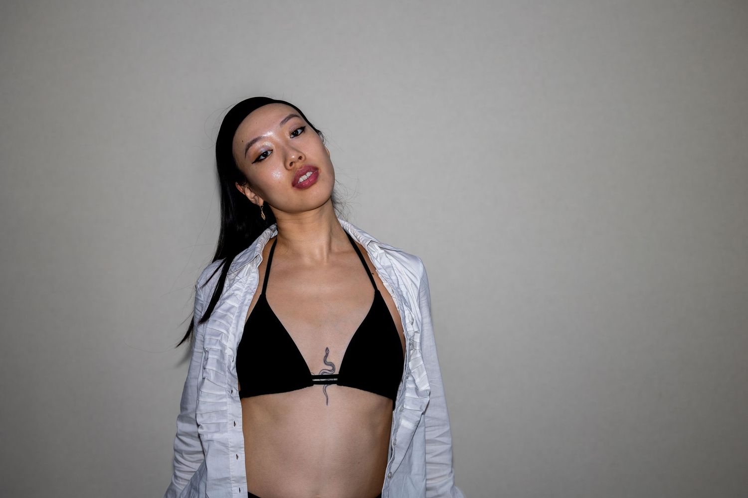 An Asian woman in a black triangle bikini top and open white jacket poses confidently indoors.