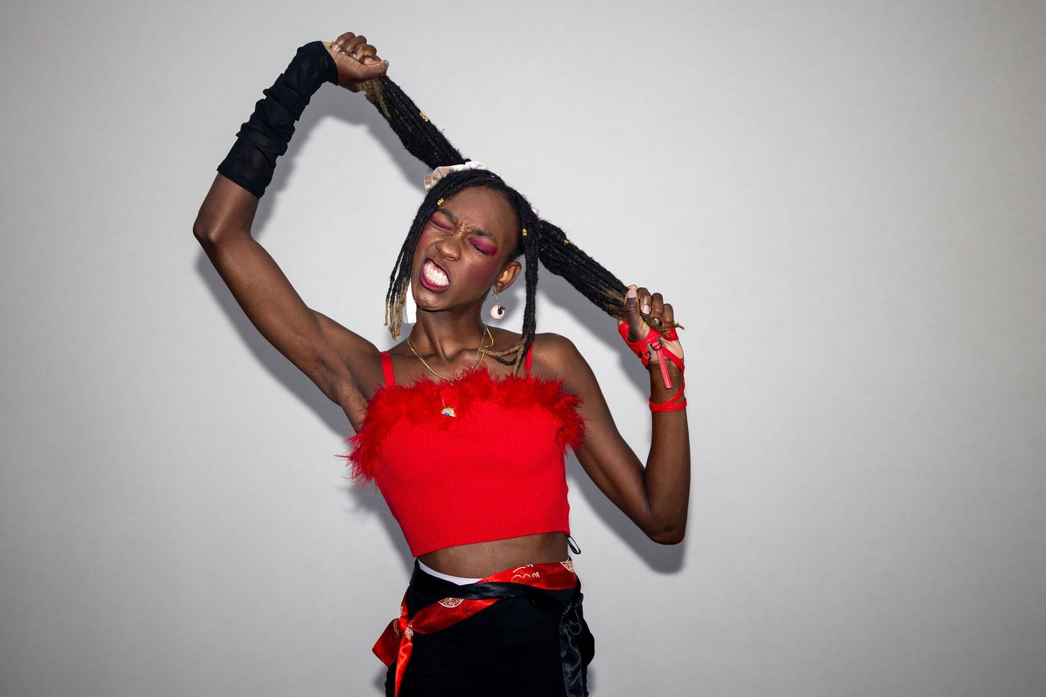 A Black woman in a red feather tube top playfully holds her long braids overhead against a light gray background.