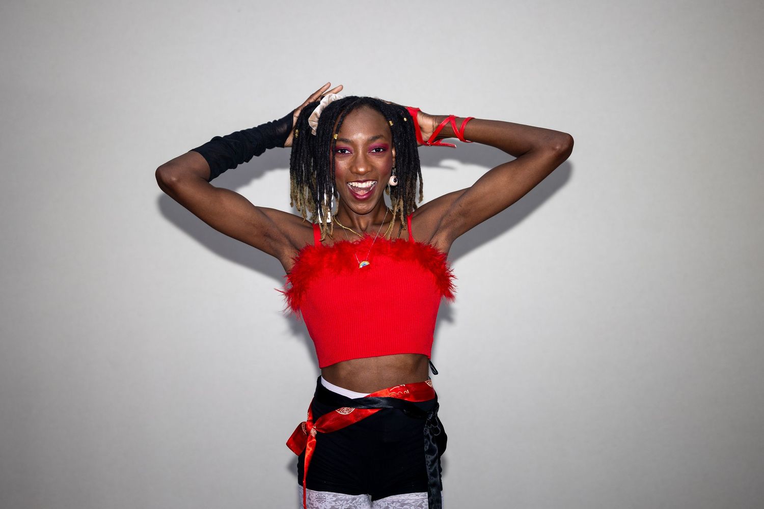 Woman in red crop top and black shorts with feather accents poses confidently with hands behind head.