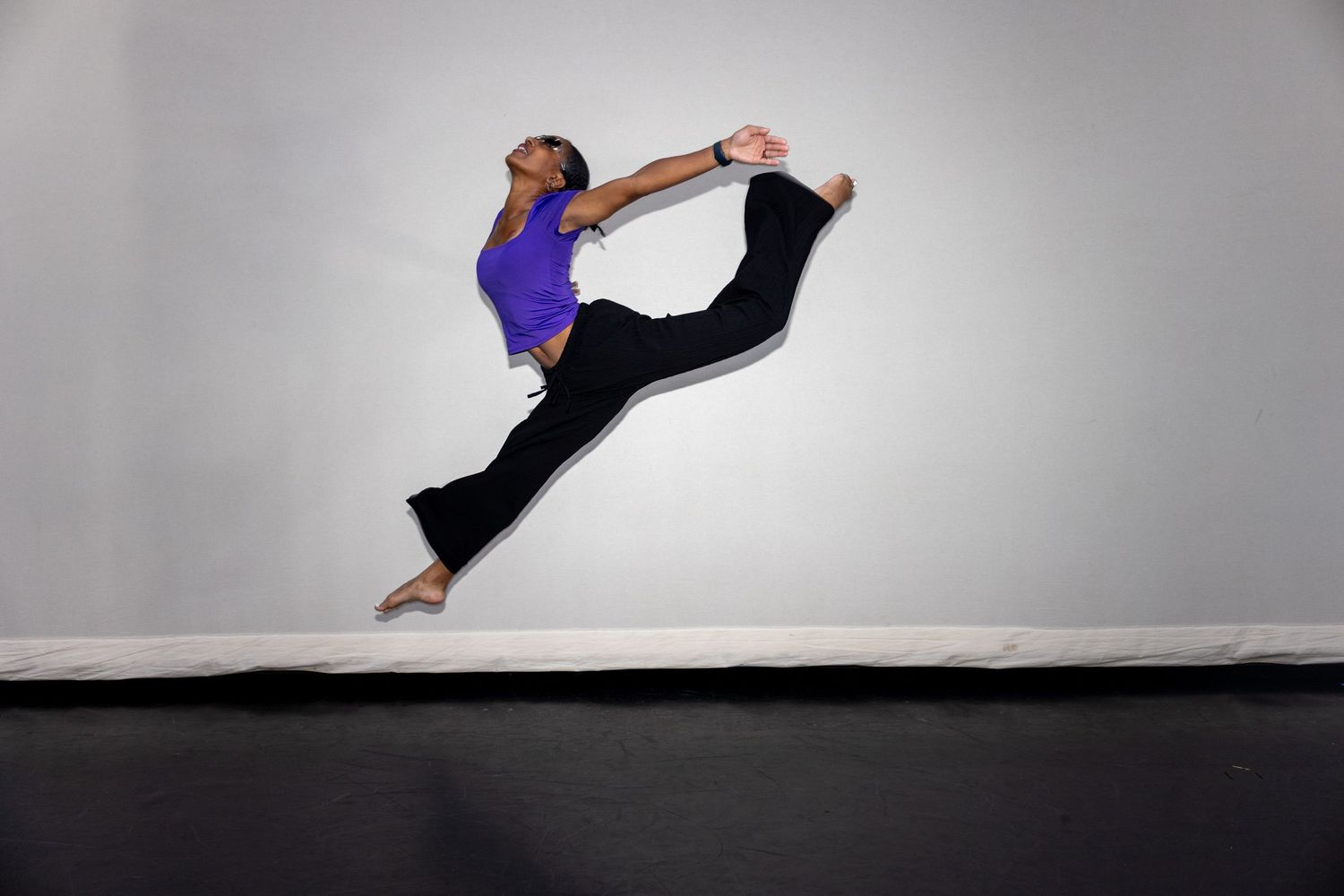 Dancer in purple top leaping mid-air with a high kick against a white studio wall.