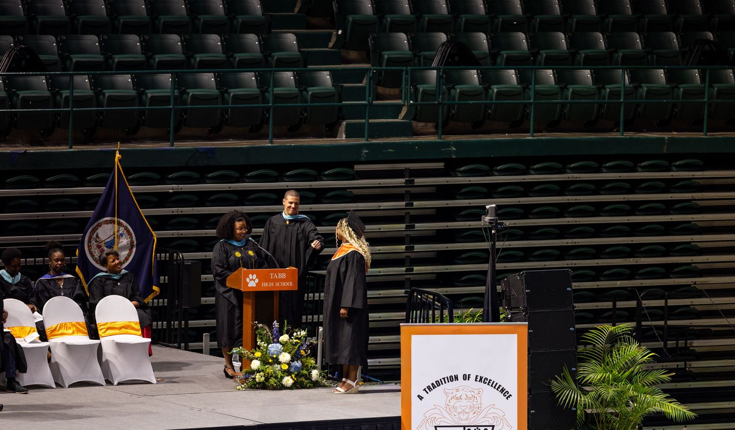 Two faculty members in academic regalia stand at a podium during a graduation ceremony in an indoor arena.