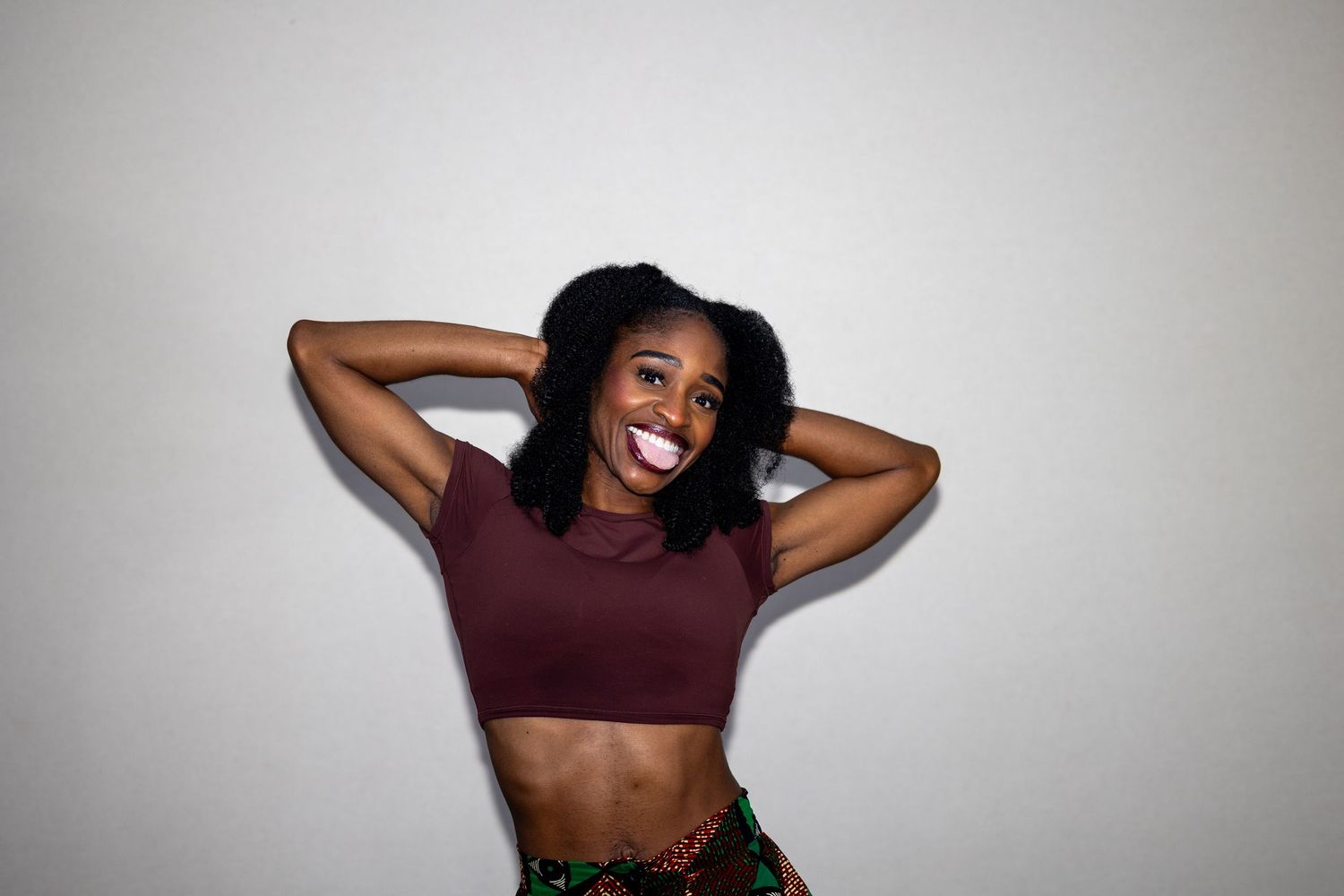 Joyful young Black woman in a maroon crop top laughing with hands behind her head against a light gray background.