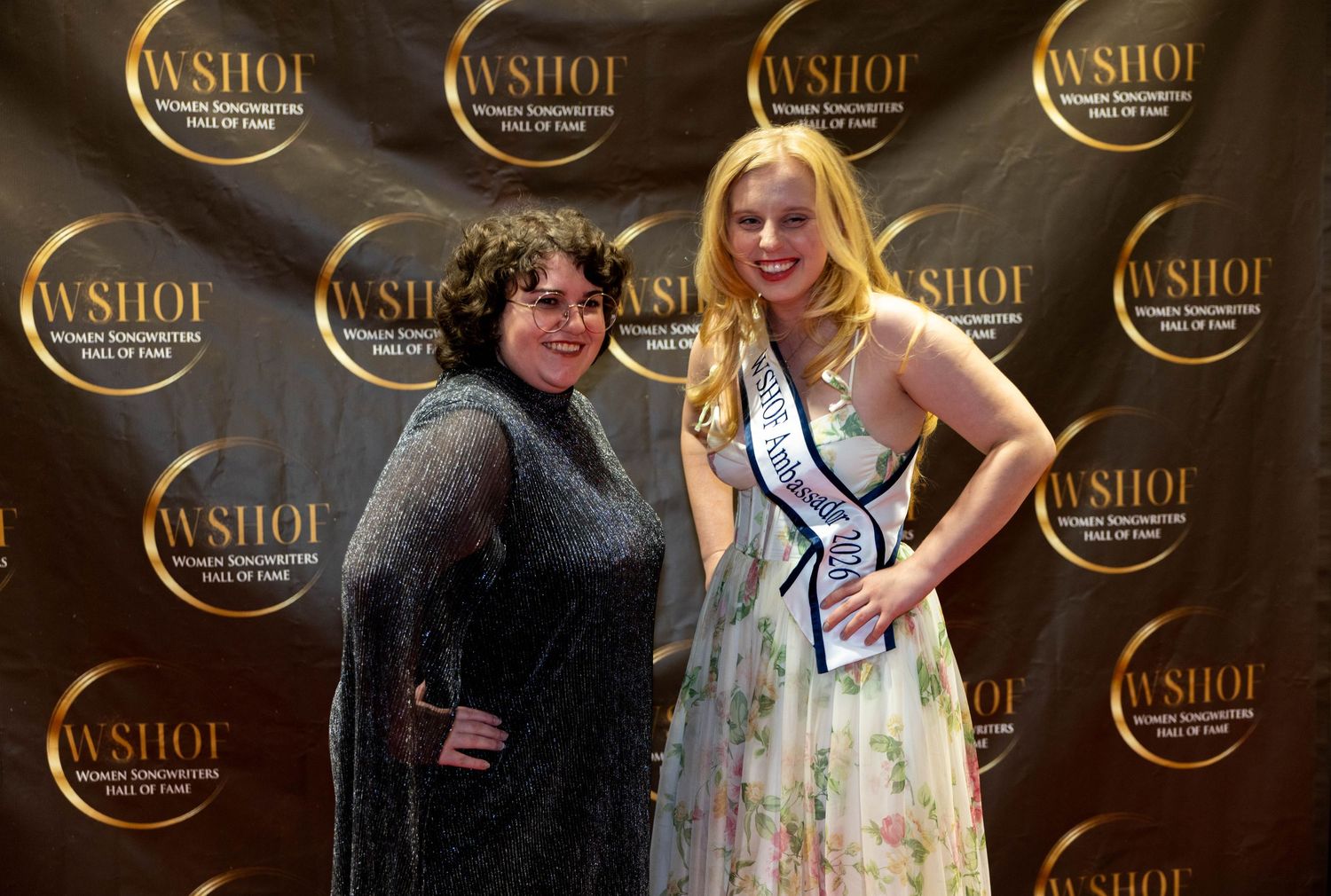 Two women smile together at WSHOF event, one wearing a pageant sash and floral gown.