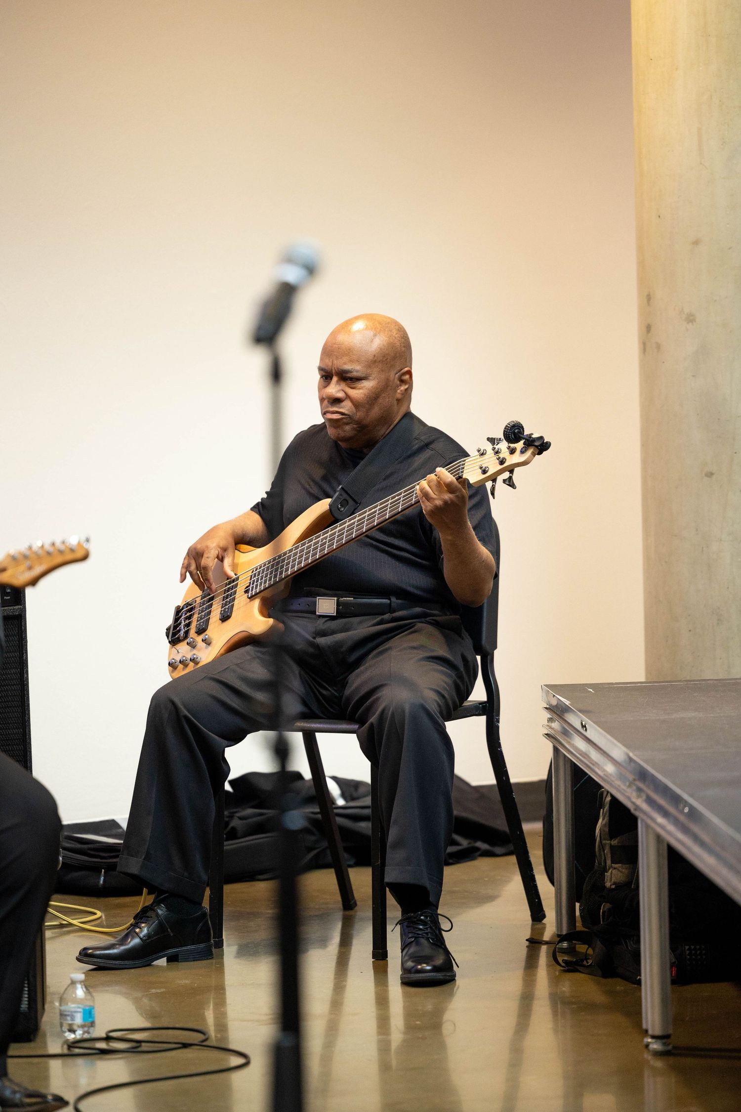 Bald man in black outfit playing bass guitar while seated on chair during indoor music performance.