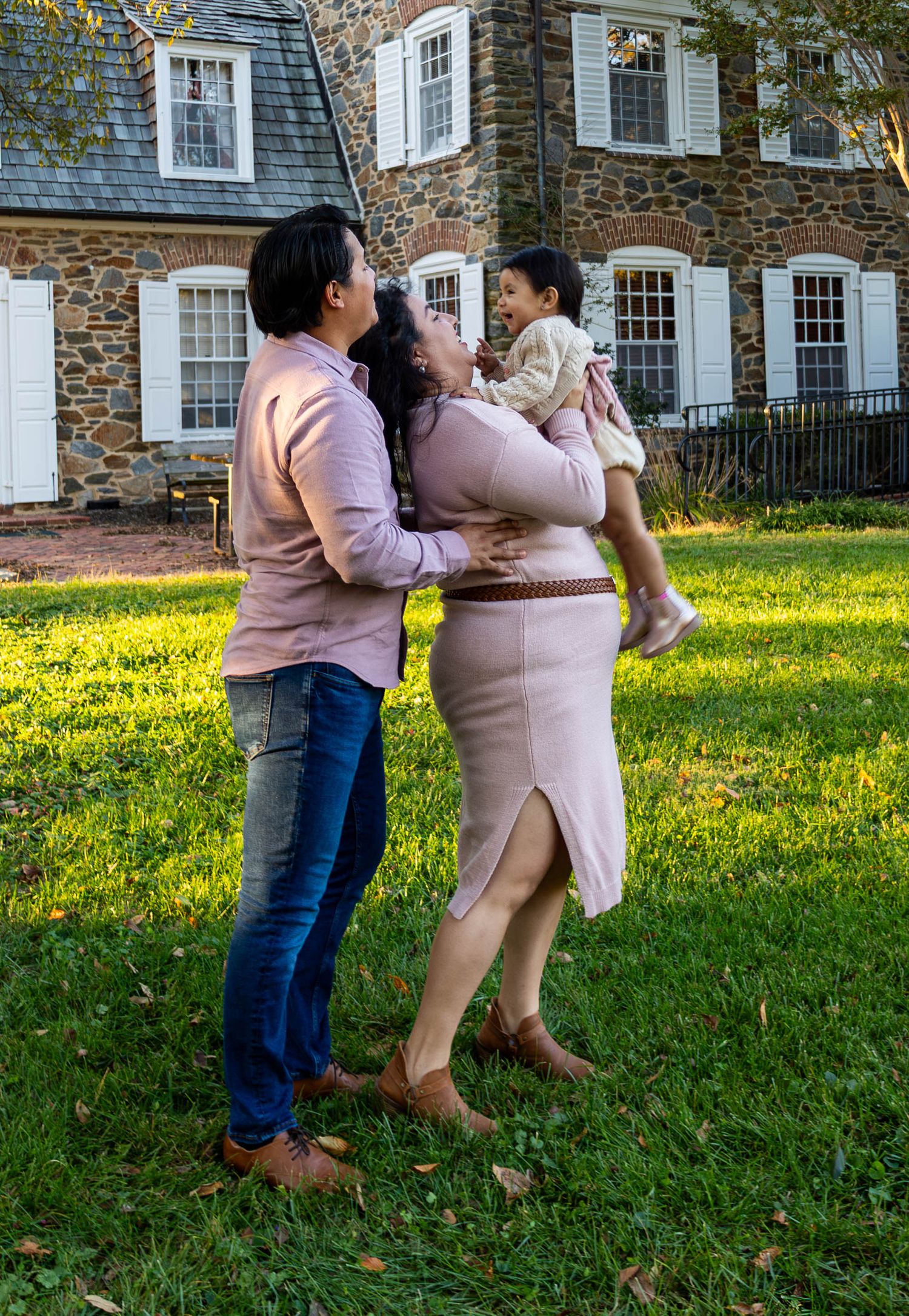 Happy family lifting toddler outdoors in front of a charming stone colonial home on green lawn.