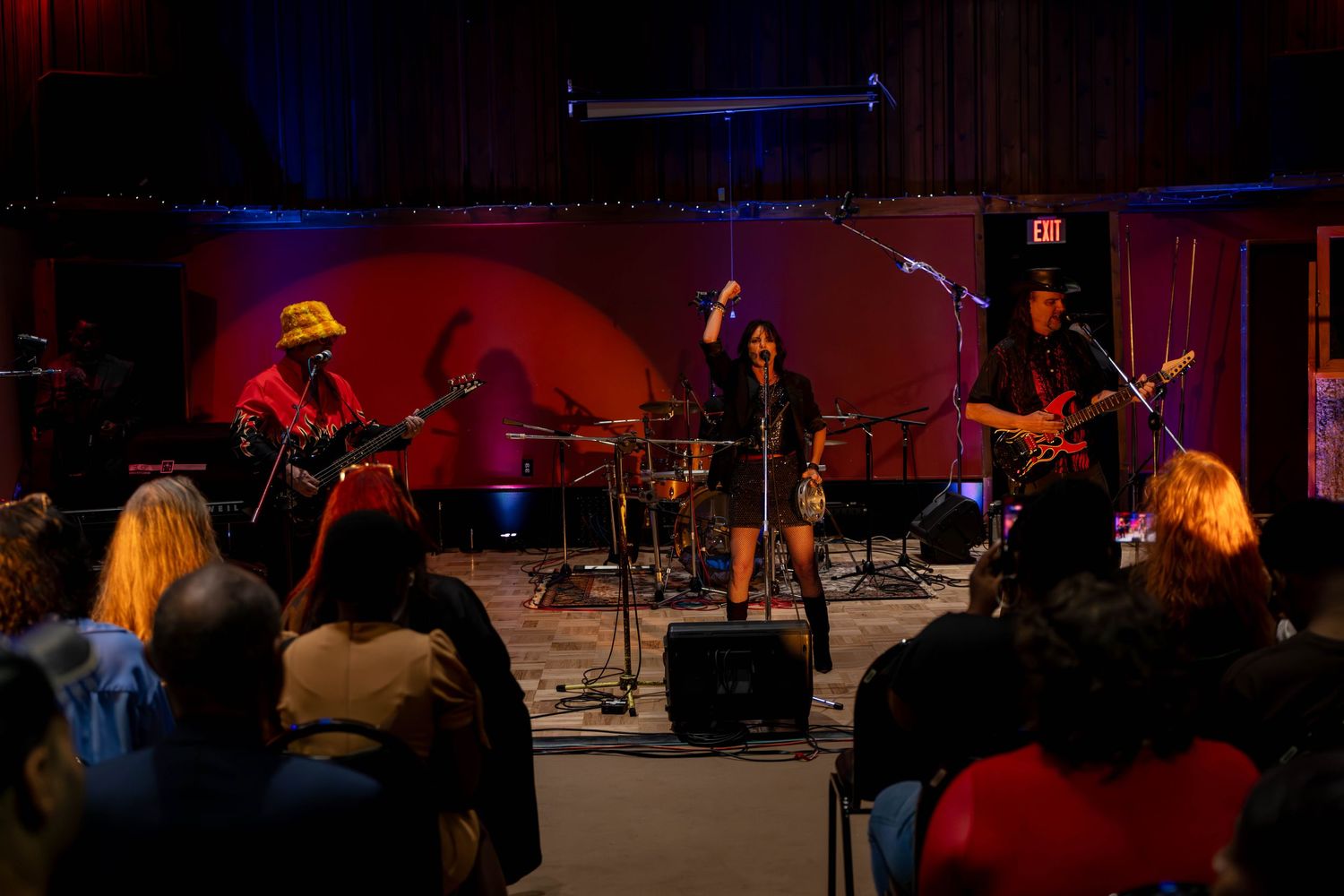 Live band performing on stage with colorful lighting, guitarist, vocalist, and drummer playing for a seated audience.