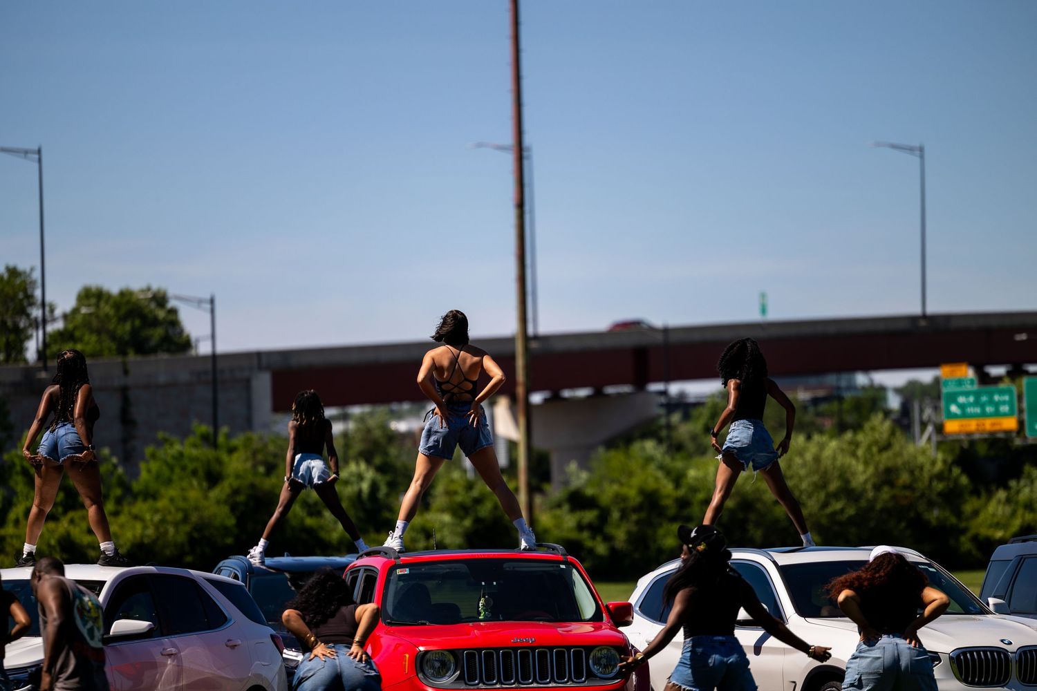 People dancing on top of cars in a sunny urban area near a highway overpass.