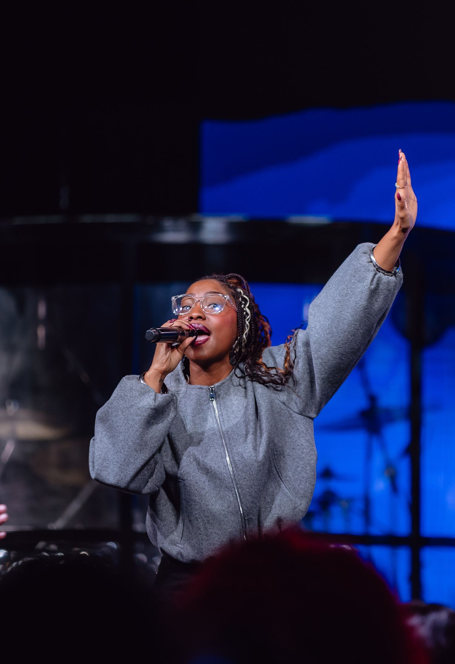 A performer in a gray sweatshirt sings passionately on stage against a dark blue background during a live performance.