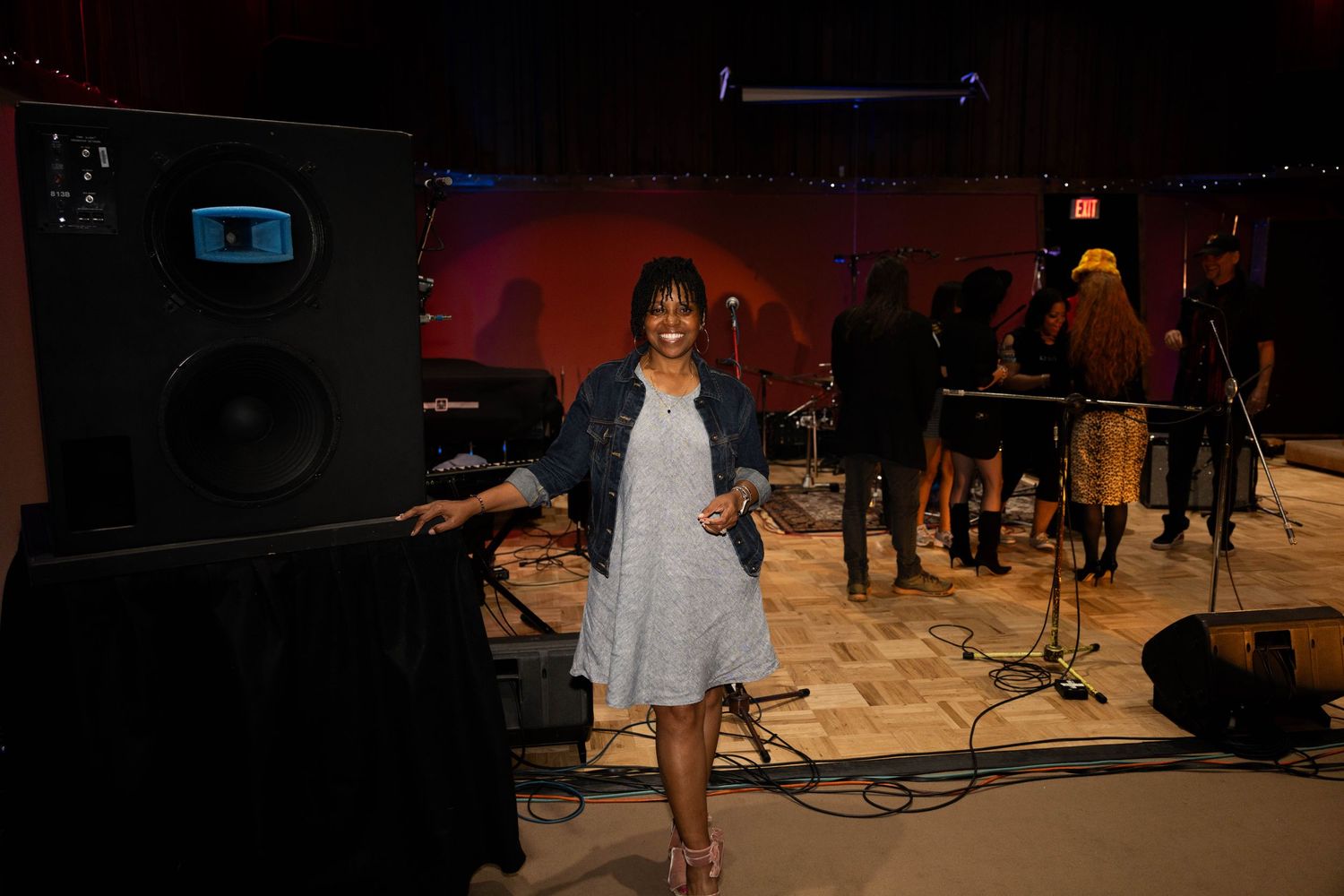 A smiling woman in a gray dress poses near a speaker in a live music venue with band members performing behind her.