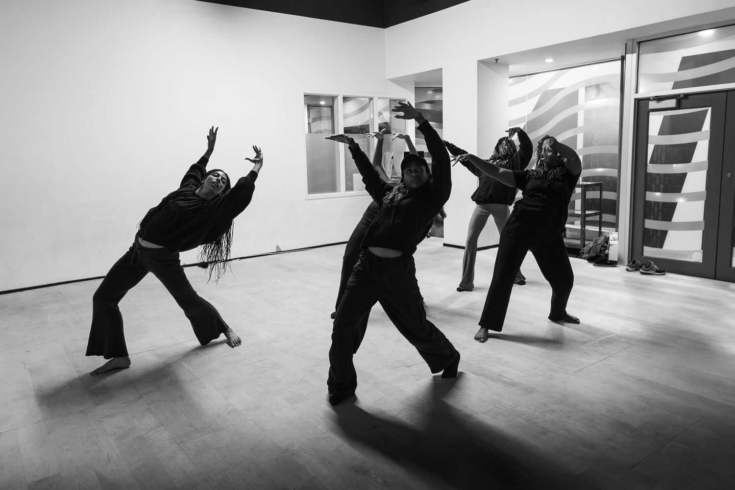 Three dancers in black attire strike dynamic poses in a minimalist studio, captured in black and white photography.