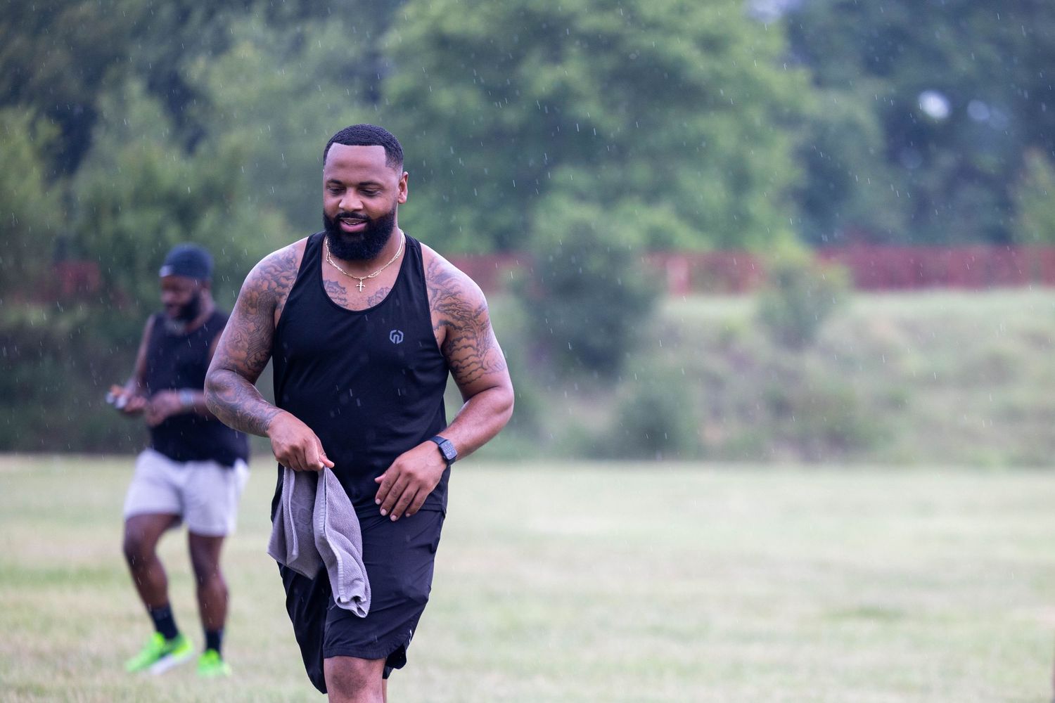 Tattooed man in black tank top smiling while jogging on green grass field during outdoor sports activity.