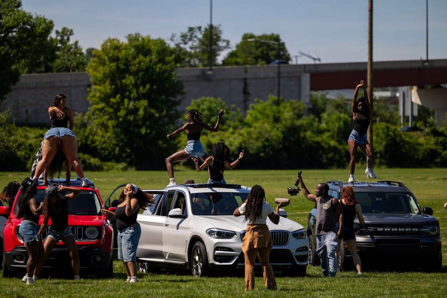 People dancing on top of cars parked on a grassy field on a sunny day.