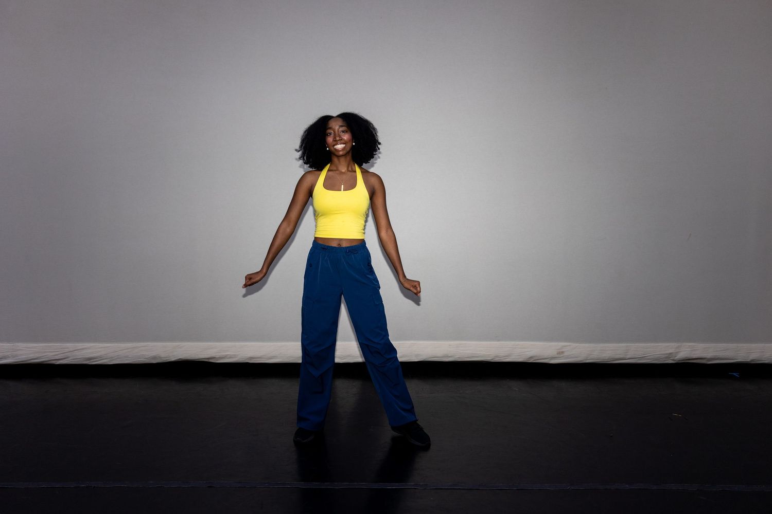 Young Black woman dancing in yellow tank top and blue wide-leg jeans against a gray studio backdrop.