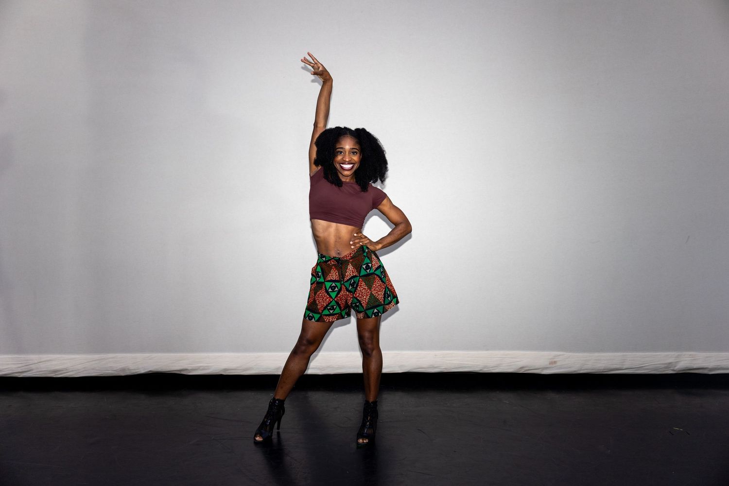 Woman in colorful African print skirt and crop top dancing with arm raised against gray background.