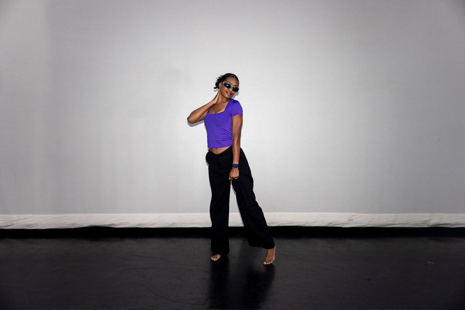 Woman in purple top and black pants posing with sunglasses on dark stage.