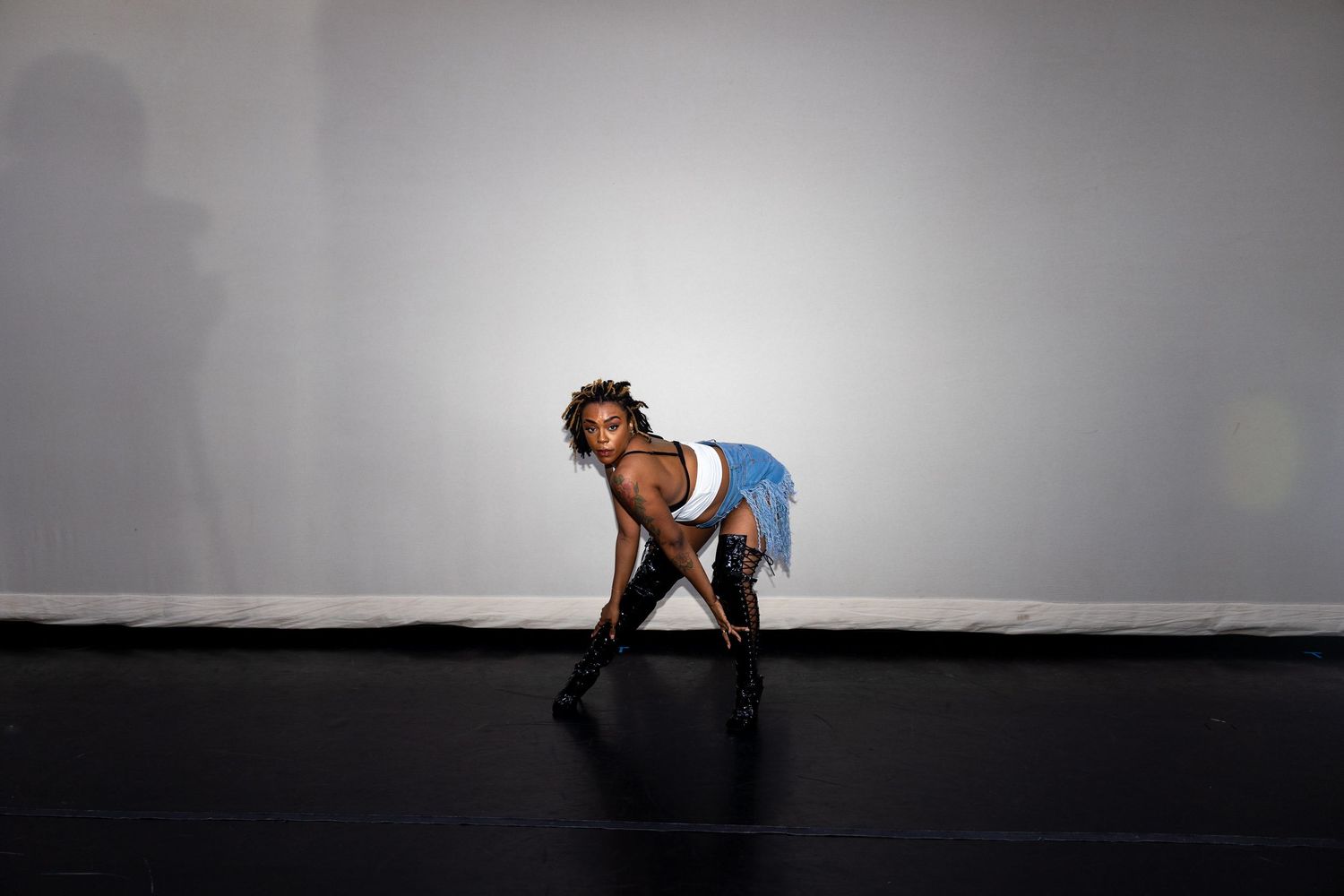 A dancer in a blue fringe skirt and boots performs a low dramatic pose on a black stage floor.