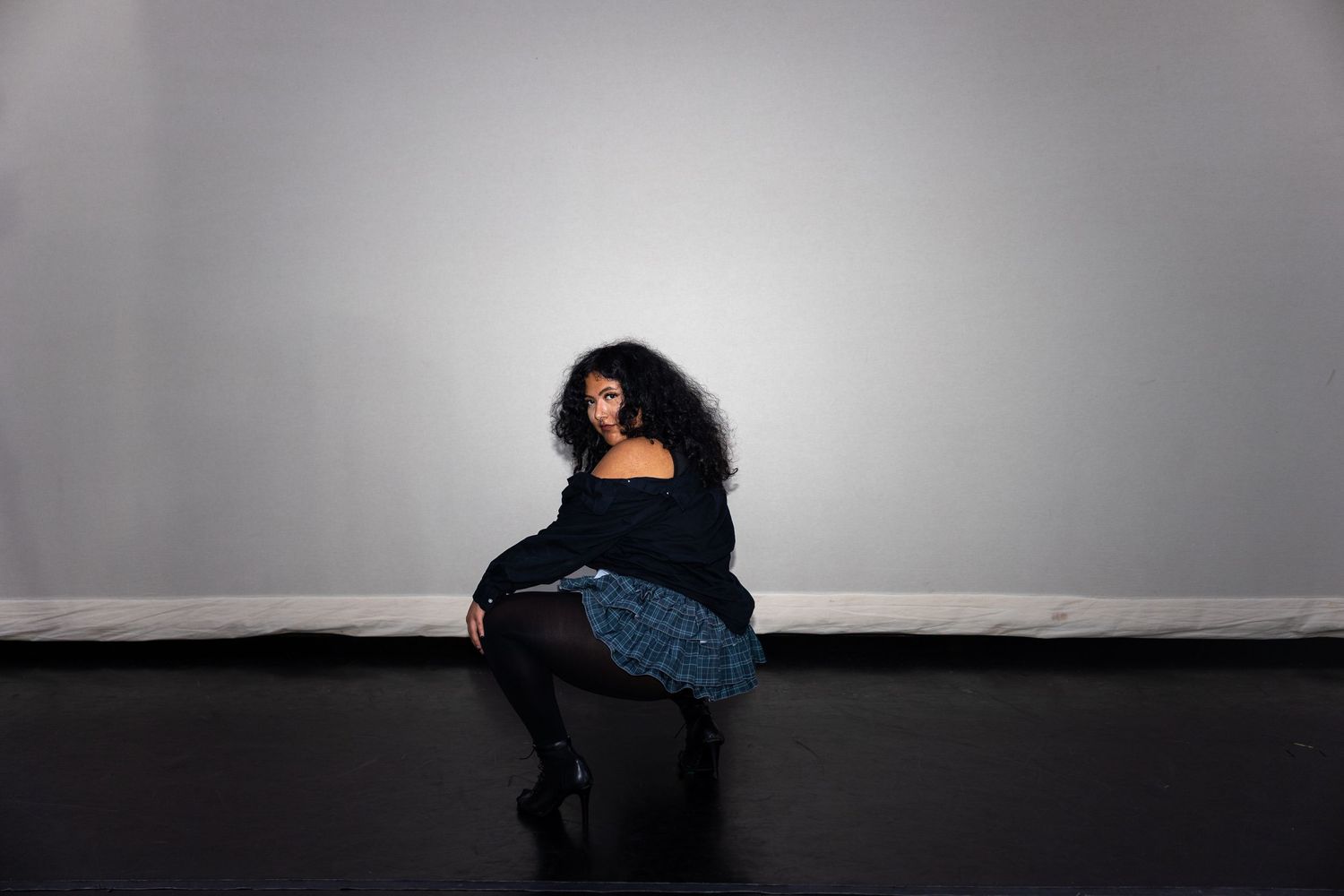 A woman with curly hair sits on a black floor against a gray wall wearing a dark off-shoulder top and plaid skirt.