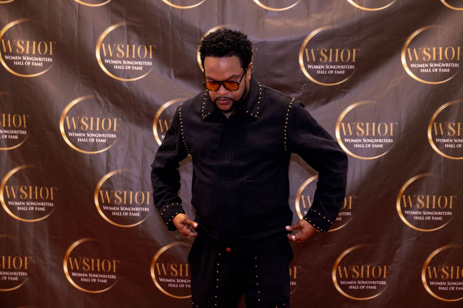 Man in black studded outfit posing at WSHOF Women Songwriters Hall of Fame event backdrop.