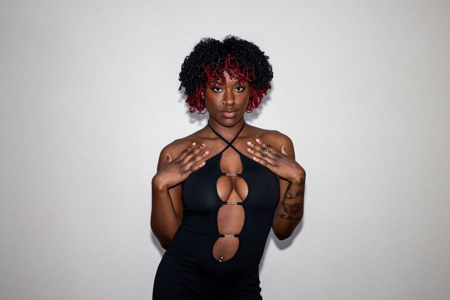 Black woman with red-tipped natural hair wearing a black cutout dress, posing with hands on chest.