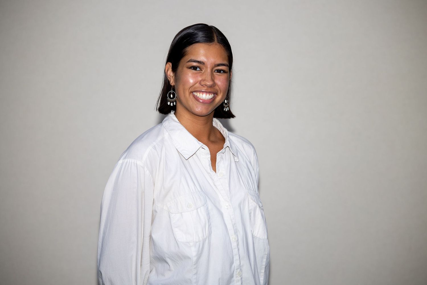 A cheerful woman wearing a white shirt and statement earrings smiles warmly against a neutral gray background.