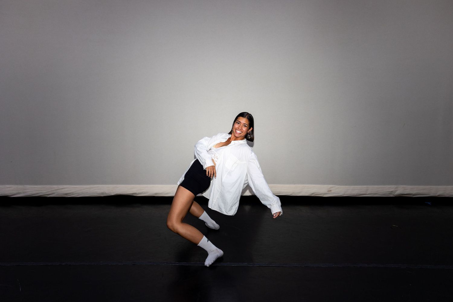 A dancer in a white oversized shirt and dark shorts strikes a dynamic pose on a black studio floor.
