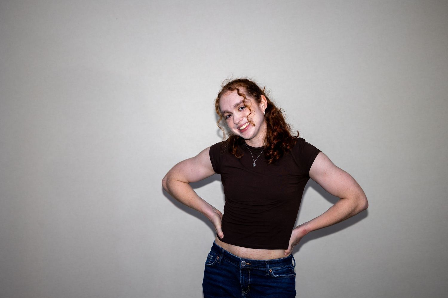 Smiling young woman with auburn hair wearing a black crop top and blue jeans posing with hands on hips.