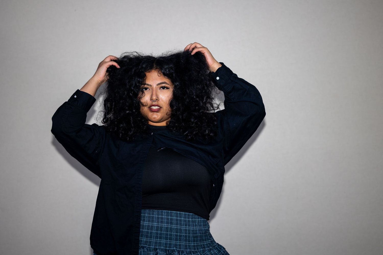 Woman with curly hair posing with hands in hair, wearing black top and plaid skirt.