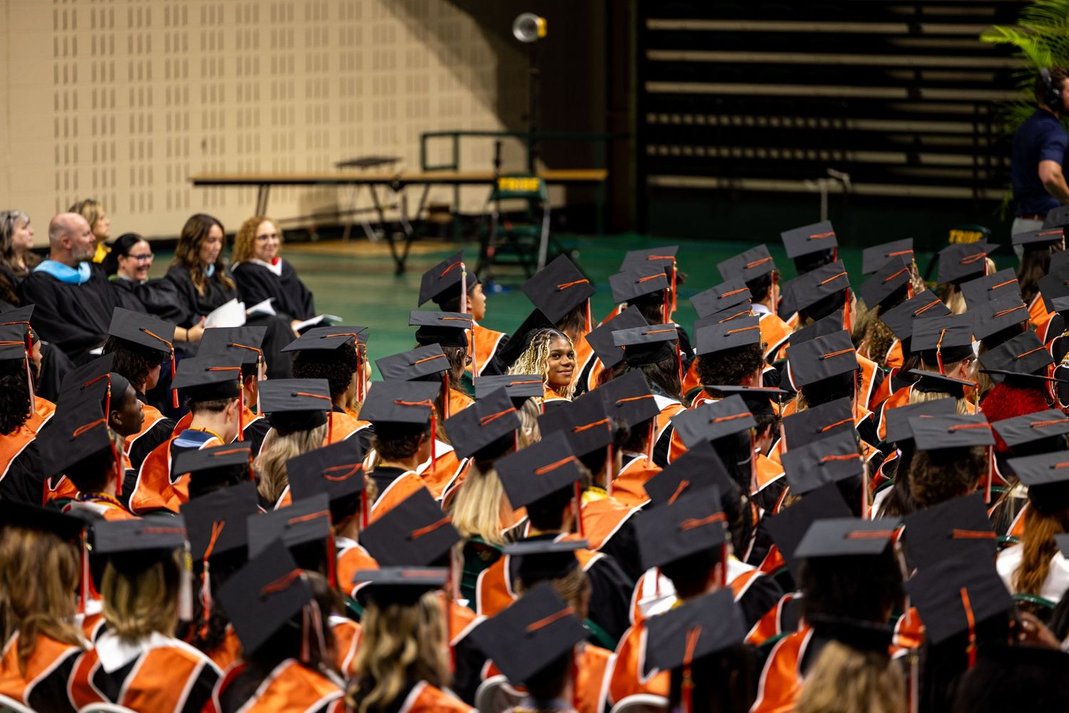 Graduates in black and orange caps and gowns seated at an indoor university commencement ceremony.