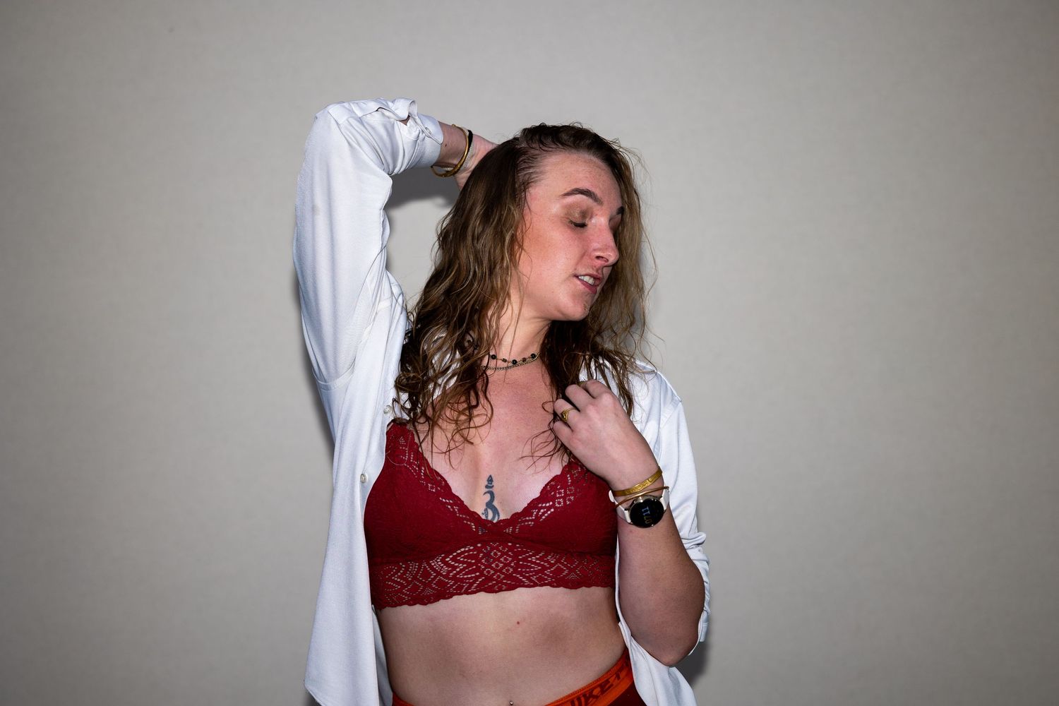 Woman with curly hair in a red lace bralette and white open shirt looking down gracefully.