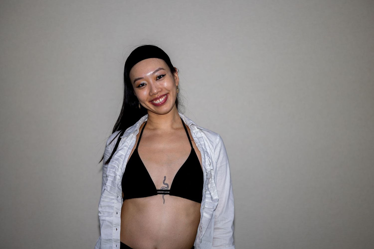 A smiling woman in a black bikini top and open white shirt shows a baby bump against a gray background.