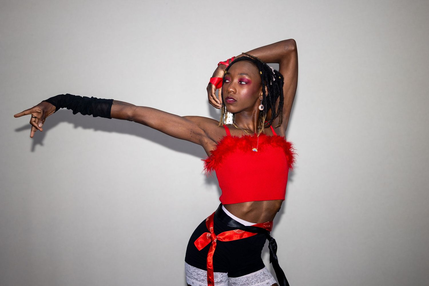A Black woman in a red feathered crop top extends her arm dramatically while wearing black gloves and a red sash.