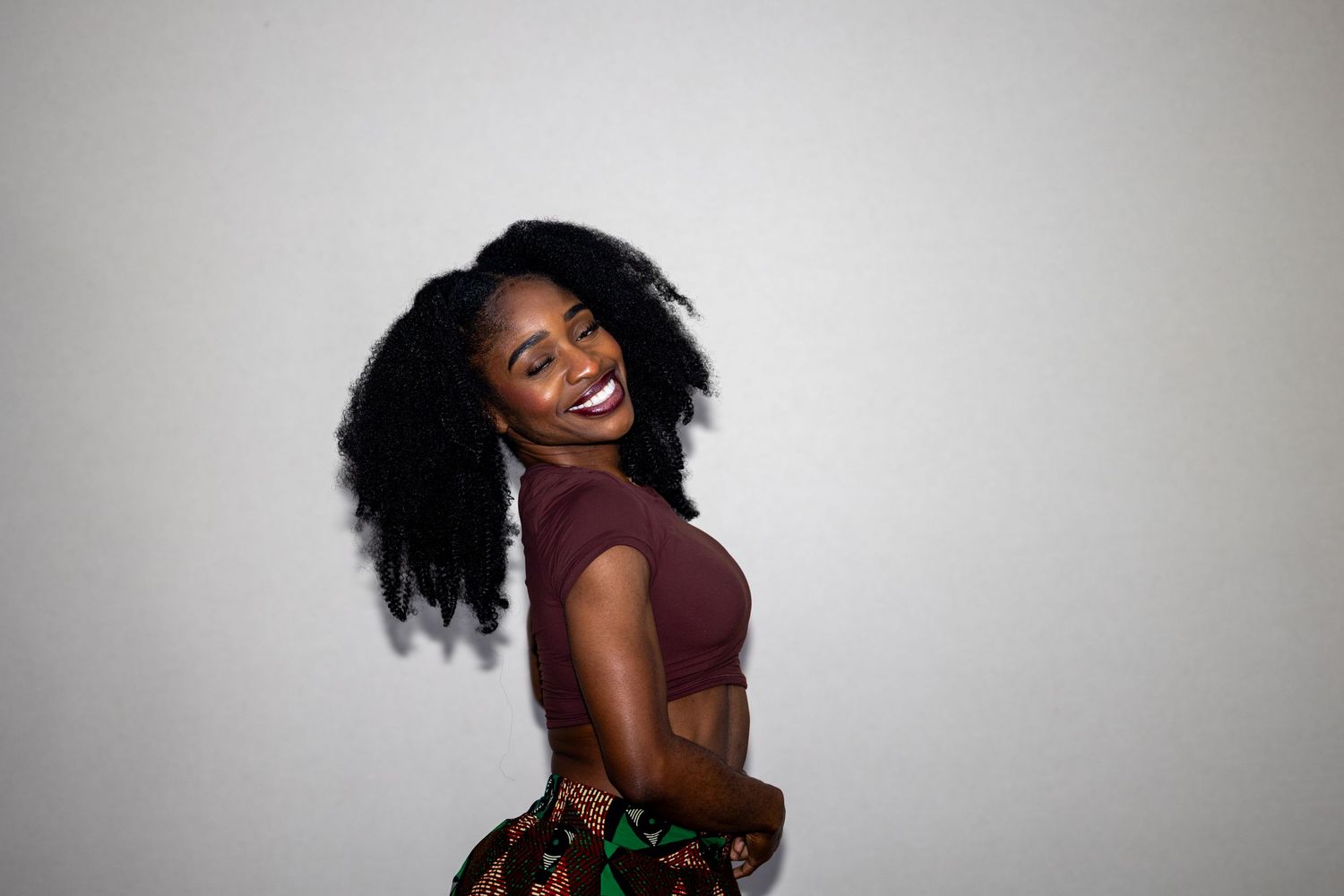 Young Black woman with natural hair in a maroon crop top posing sideways and smiling against a white background.