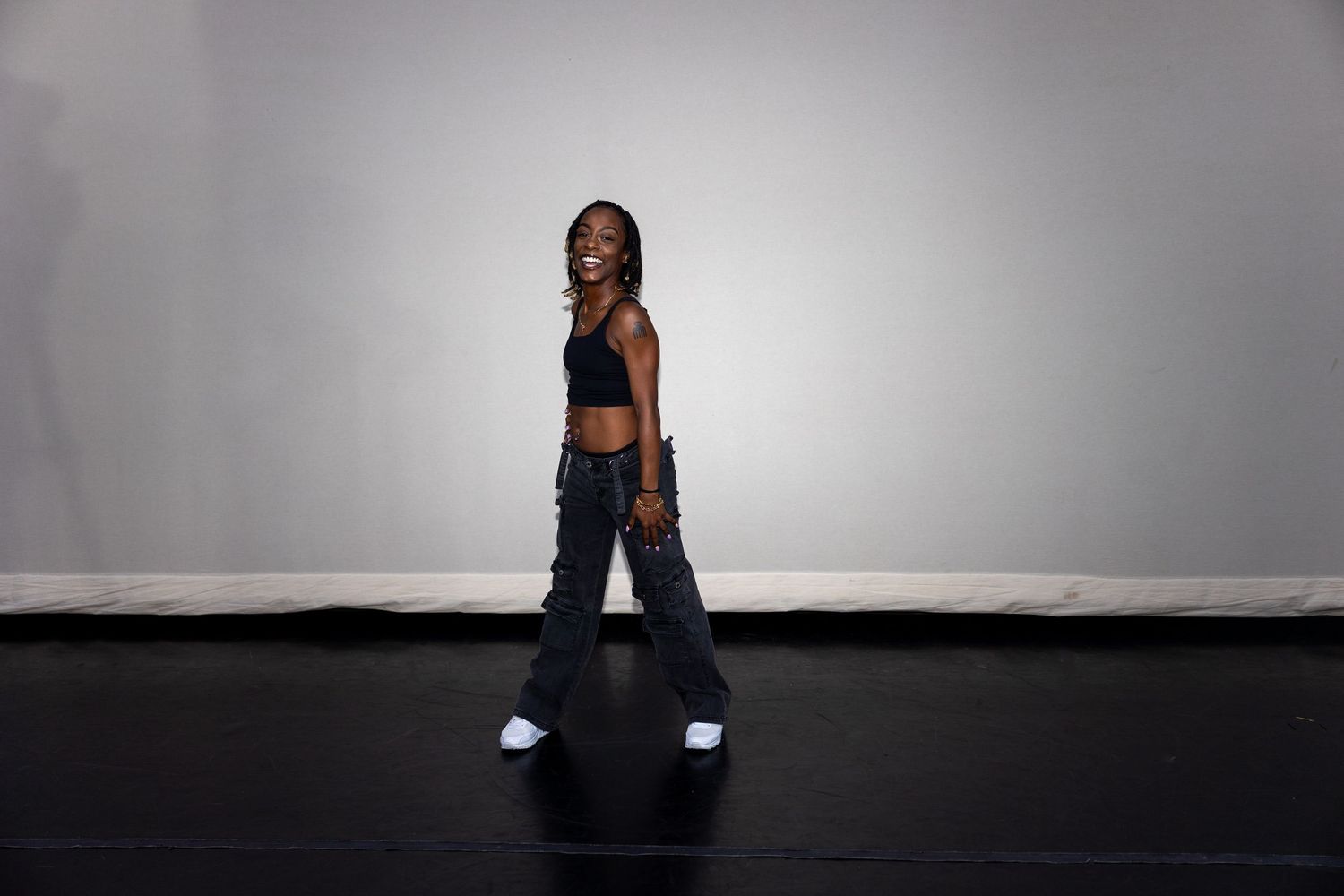 Black woman in black crop top and wide-leg cargo pants walks across dark studio floor.