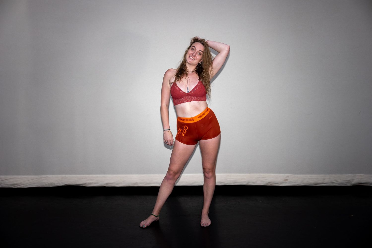 A smiling dancer poses in a red tie-dye sports bra and matching shorts against a white studio wall.