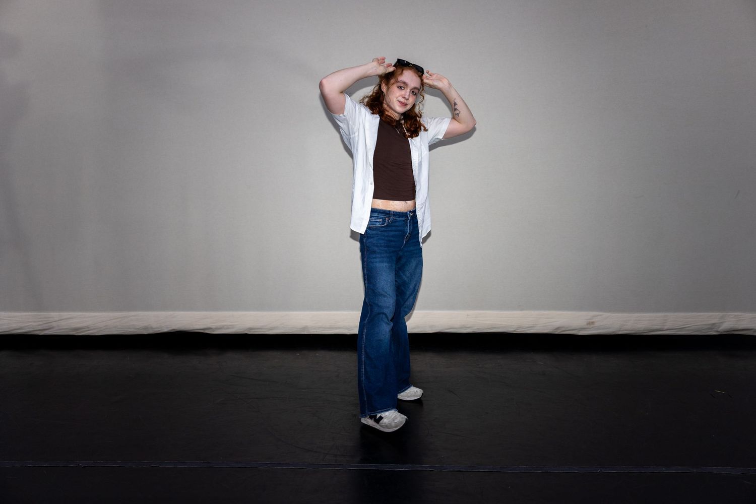 A dancer poses in wide-leg jeans, brown crop top, and white shirt with hands on head in a studio.