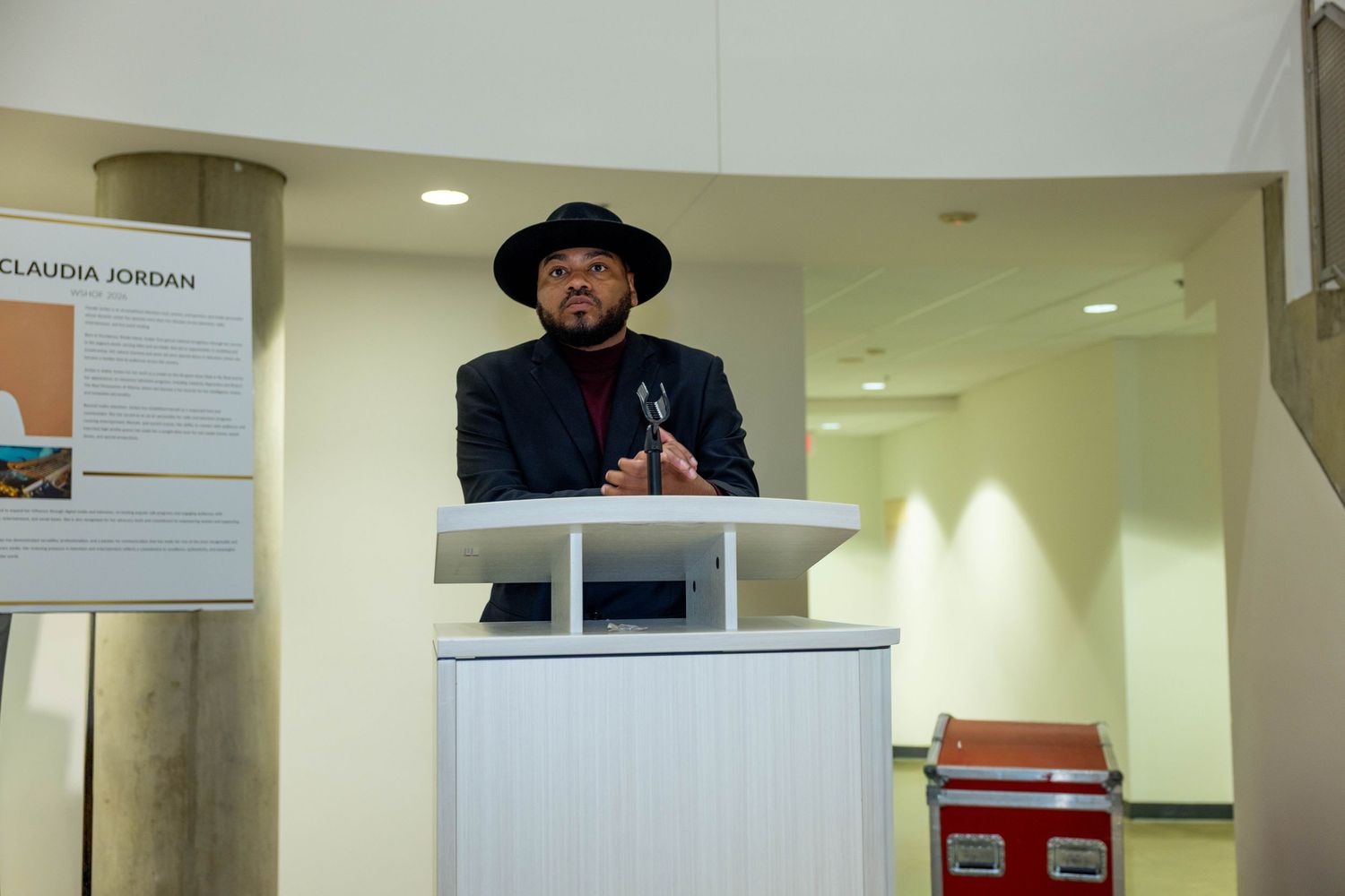 Man in black hat and jacket speaking at a white podium during an indoor event or ceremony.