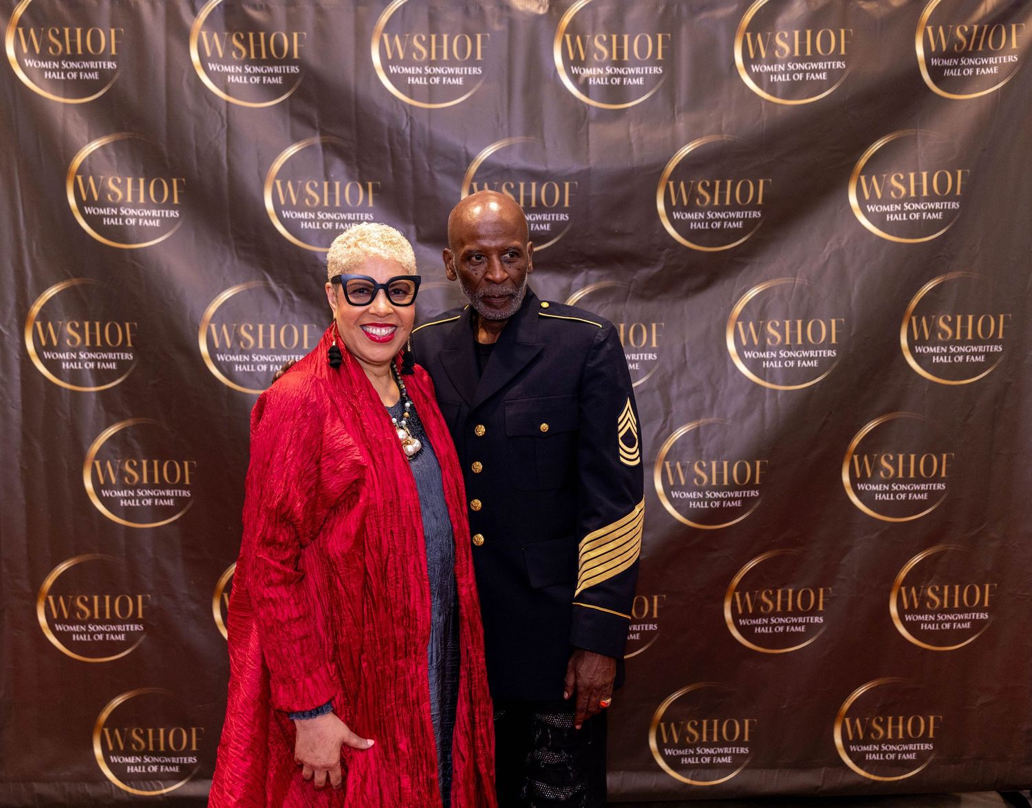 A woman in a red jacket and a man in a military uniform pose together in front of a WSHOF step-and-repeat backdrop.