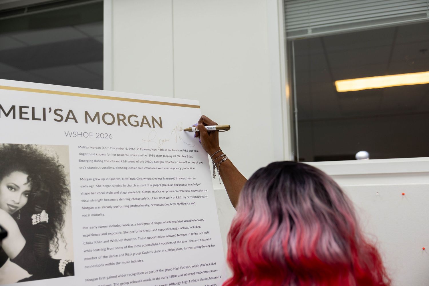 Person with red hair signing a Meli'sa Morgan VSHOW 2016 promotional poster at an event.