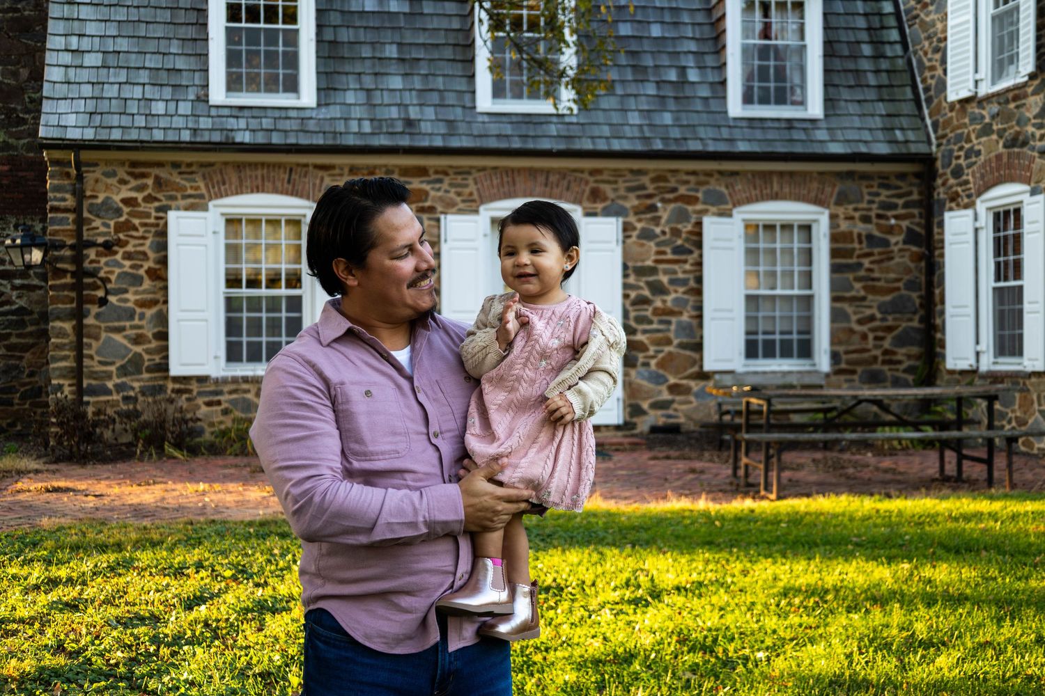 Man holding smiling baby girl in pink dress outside historic stone house on green lawn.